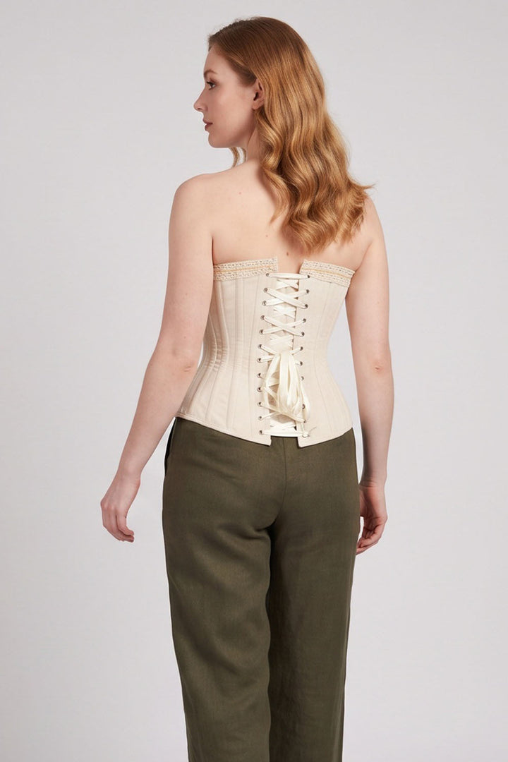 A woman shown from the back wearing a Luxe Noir cream-colored strapless corset top with lace-up detailing and olive green trousers, set against a plain light gray studio background.