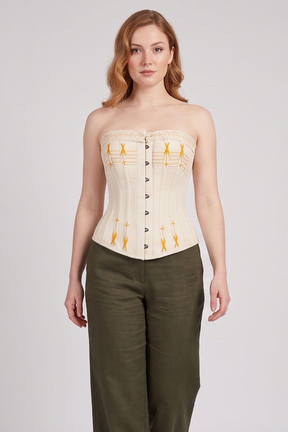 A woman modeling a strapless Luxe Noir corset top in cream with yellow geometric patterns and dark buttons, paired with olive green pants, against a light gray studio background.