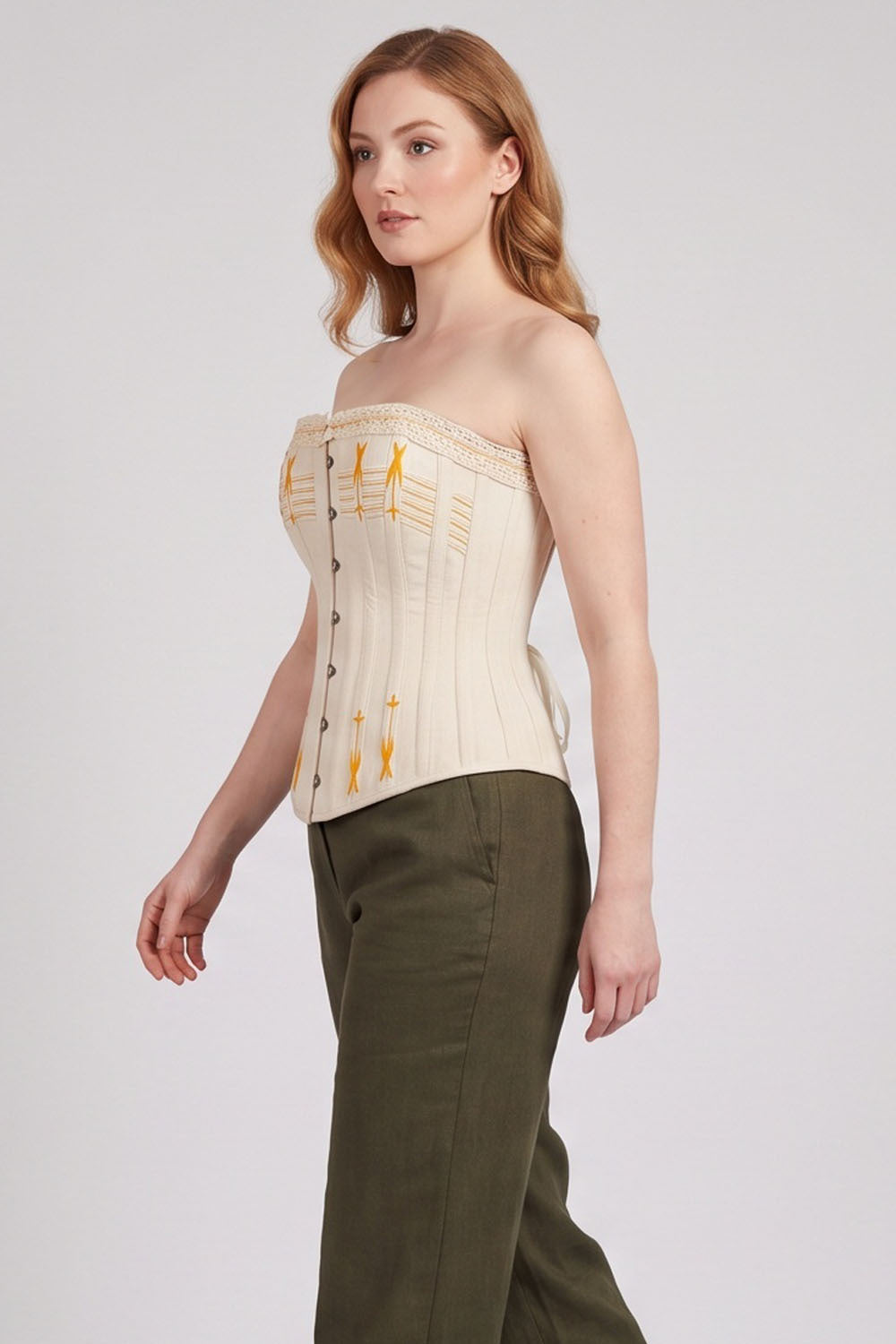 A woman modeling a strapless Luxe Noir cream corset-style top with gold embroidered details and olive green trousers, photographed against a light gray studio background.