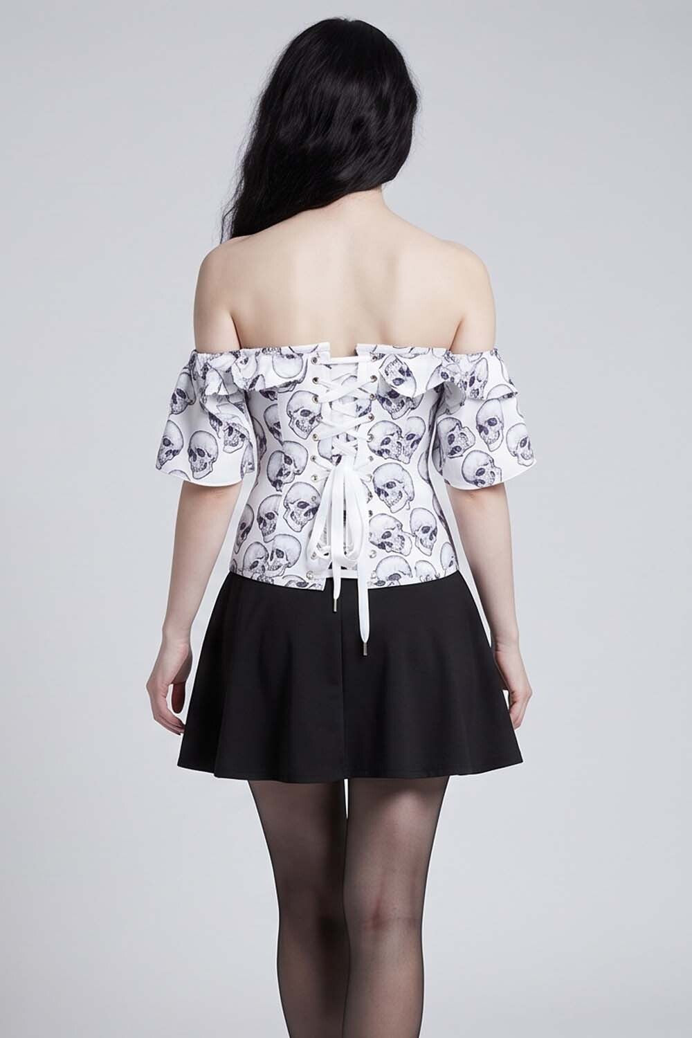 A model shown from the back wearing an Luxe Noir Corset off-the-shoulder white top with a black skull pattern and a black pleated skirt, set against a plain light gray studio background.