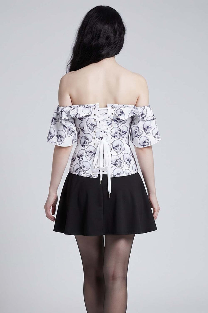 A model shown from the back wearing an Luxe Noir Corset off-the-shoulder white top with a black skull pattern and a black pleated skirt, set against a plain light gray studio background.