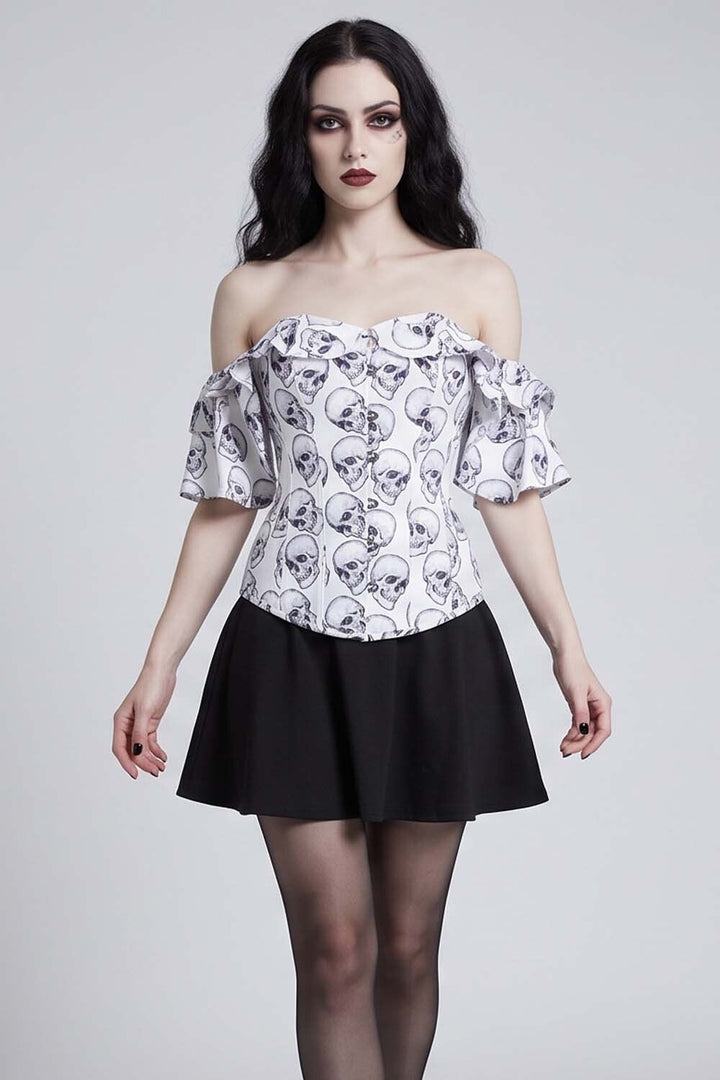 A woman modeling an Luxe Noir Corset off-the-shoulder white top with a black skull and crossbones pattern, paired with a black skirt, against a light gray studio background.