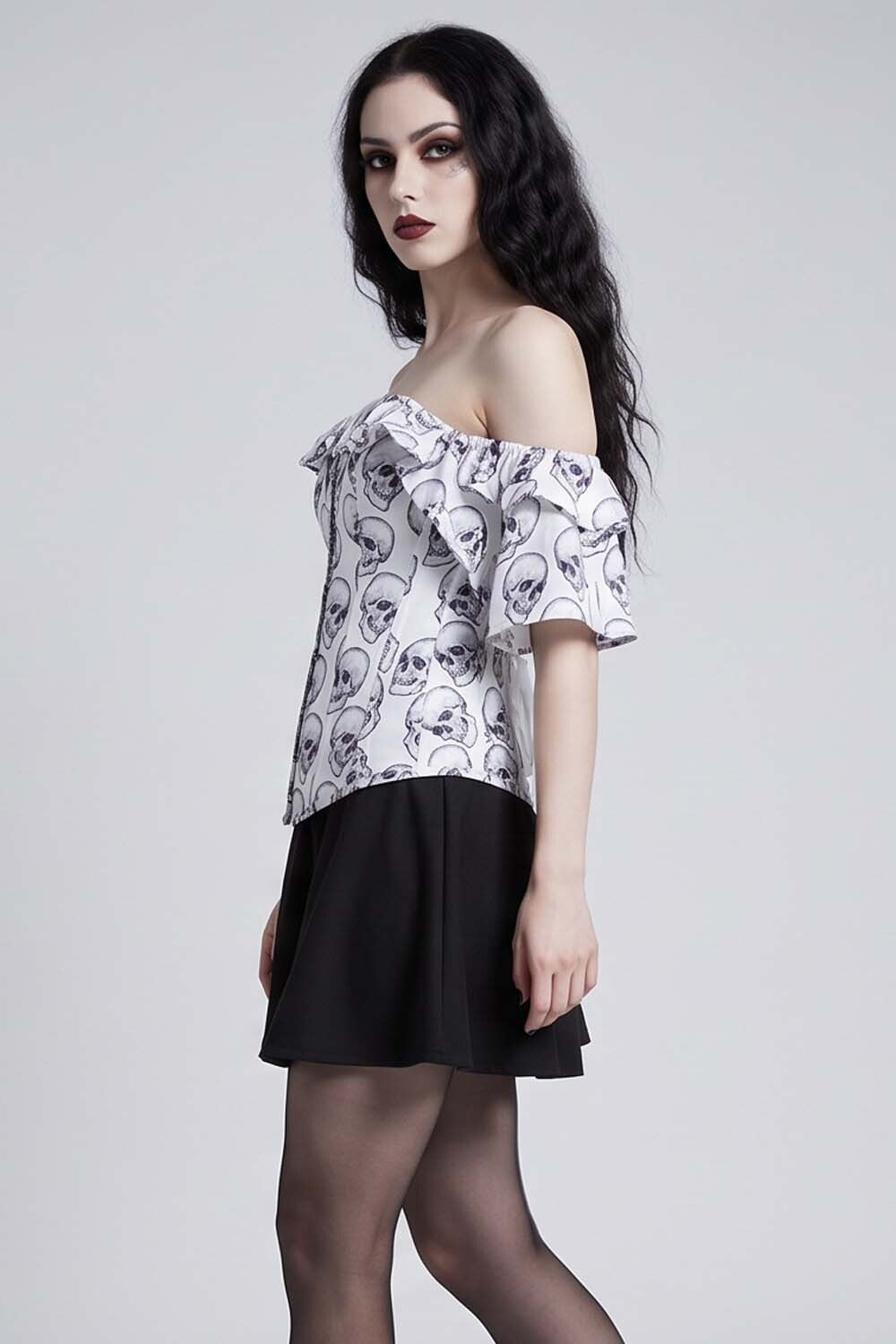 A woman modeling an Luxe Noir Corset off-the-shoulder blouse with a white base and black skull pattern, paired with a black skirt and sheer black tights, against a light gray studio background.