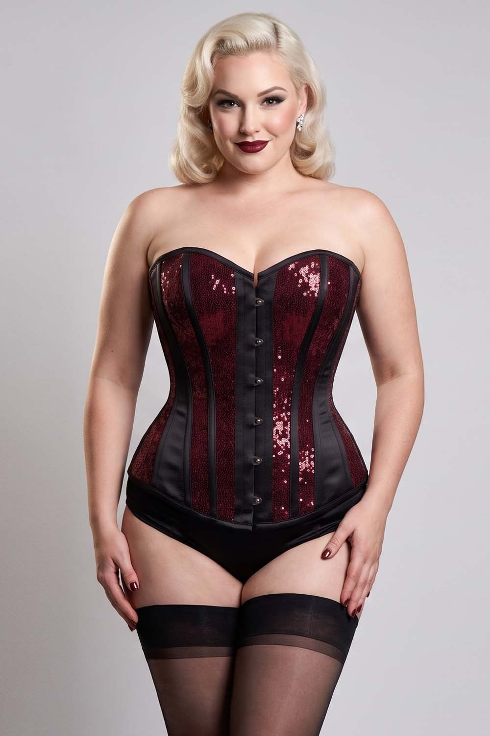 A plus-size blonde model wearing a strapless corset with deep red sequins and black panels, paired with black underwear and thigh-high stockings, against a light gray studio background.