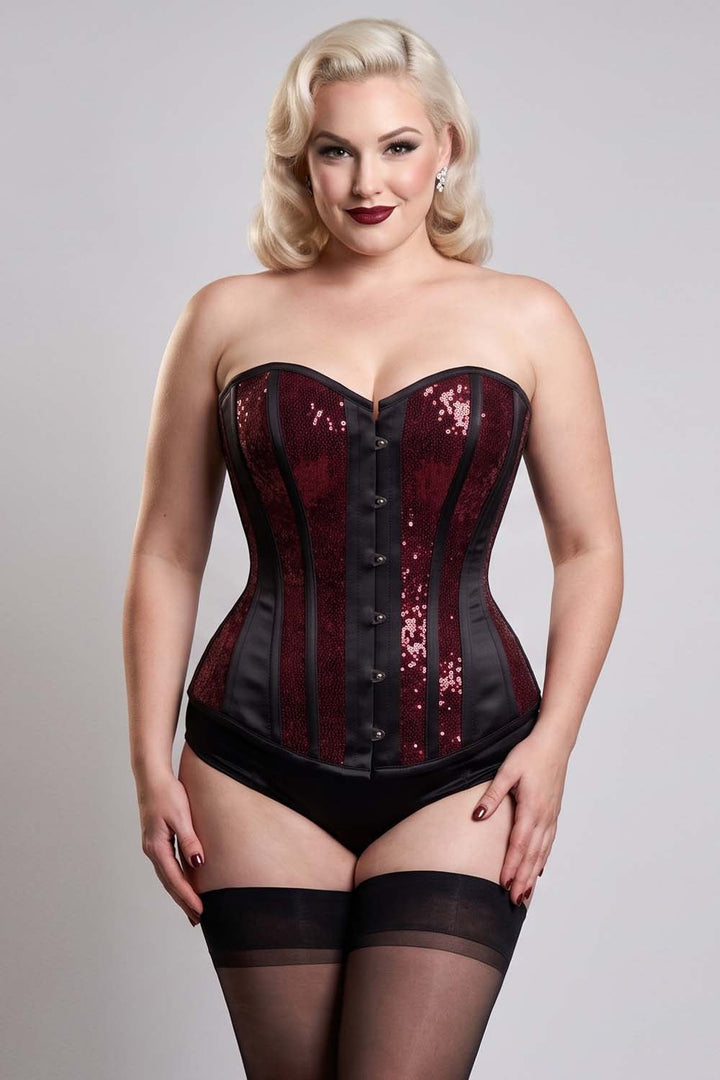 A plus-size blonde model wearing a strapless corset with deep red sequins and black panels, paired with black underwear and thigh-high stockings, against a light gray studio background.