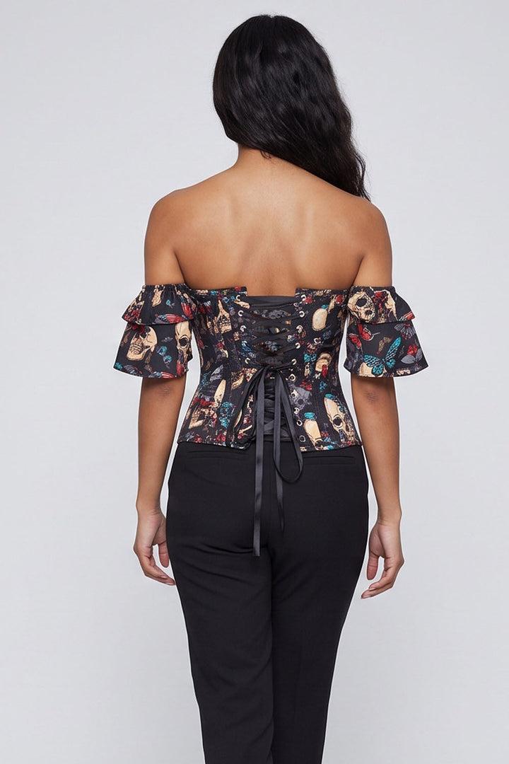 A woman shown from the back wearing a Luxe Noir  black off-the-shoulder corset top with a colorful skull and floral pattern, paired with black pants, against a light gray studio background.