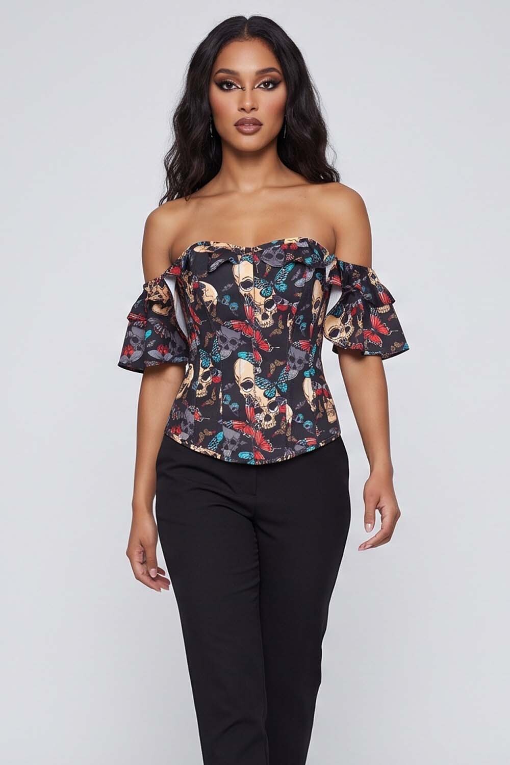 A woman modeling an Luxe Noir off-the-shoulder ruffled top with a colorful skull and floral pattern on a dark background, paired with black pants, against a light gray studio backdrop.