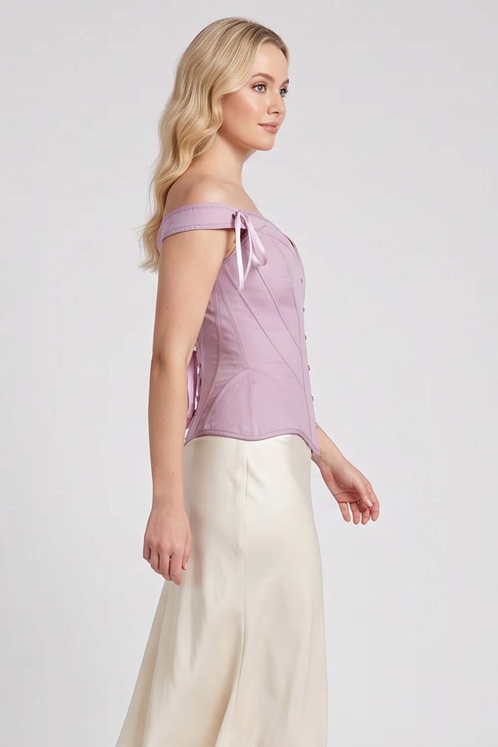 A woman modeling a lavender Luxe Noir Corset off-the-shoulder top with a corset-style bodice and cream satin skirt against a light gray studio background.