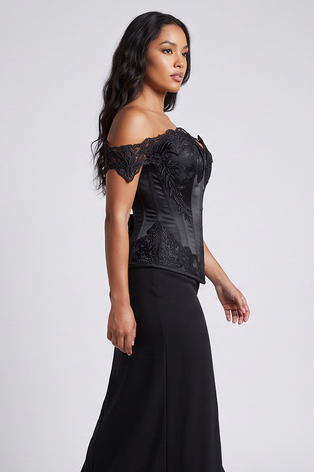 A woman modeling a Luxe Noir black off-the-shoulder evening gown with a structured lace and satin bodice and a flowing skirt, photographed against a light gray studio background.