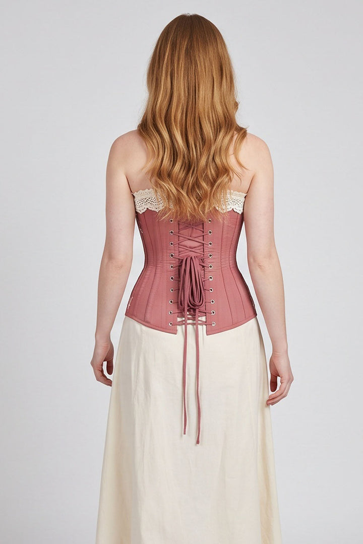 A woman viewed from the back wearing a Luxe Noir dusty rose corset with lace-up back detailing and a cream skirt, set against a plain light gray studio background.