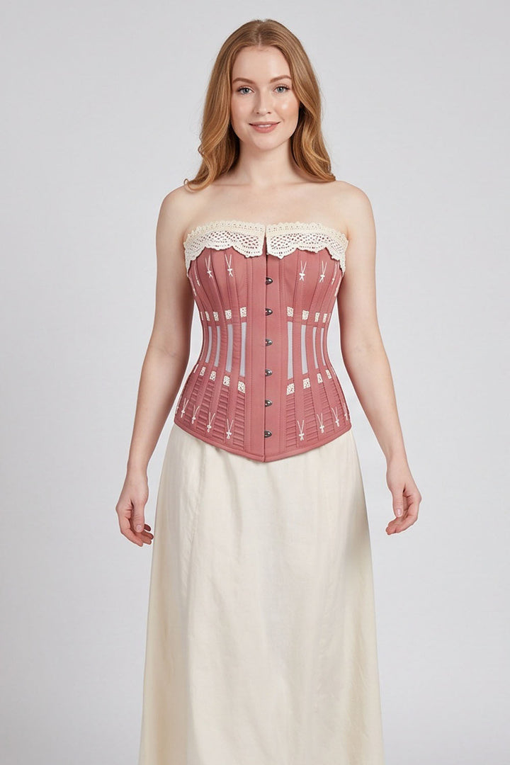A woman modeling a Luxe Noir strapless corset-style top in dusty rose pink with cream lace trim and a cream skirt against a light gray studio background.