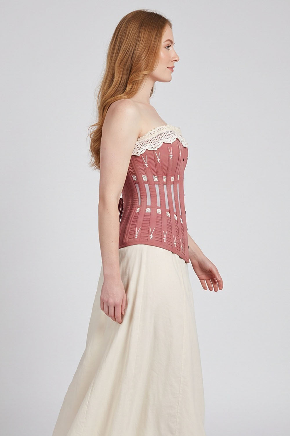 A woman in profile wearing a Luxe Noir dusty rose corset-style top with cream lace trim and a long cream skirt, photographed against a light gray studio background.