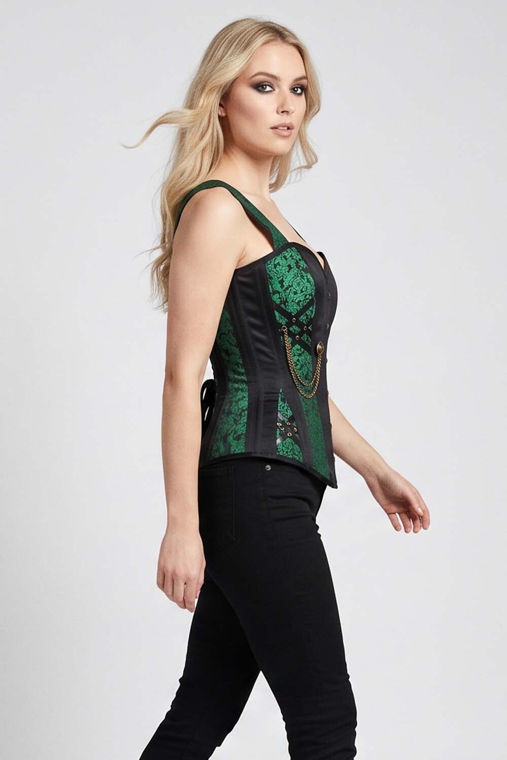 A blonde model wearing a Luxe Noir  green and black lace corset top with black pants, shown from a side profile against a light gray studio background.