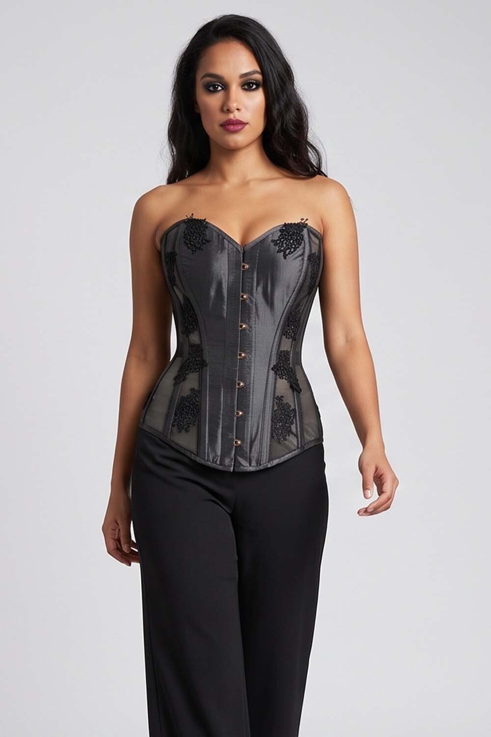 A woman modeling a Luxe Noir  strapless black lace and mesh corset top with gold-tone buttons, paired with black pants, against a light gray studio background.