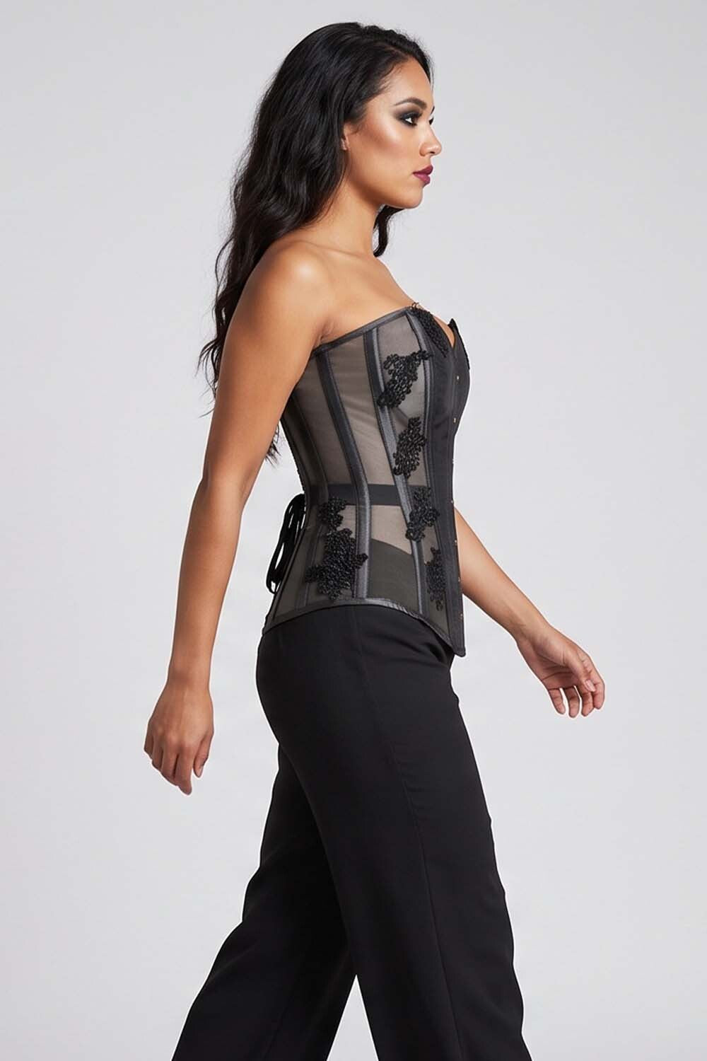 A woman modeling a black Luxe Noir   strapless corset top with sheer mesh panels and black lace appliques, paired with black pants, against a light gray studio background.