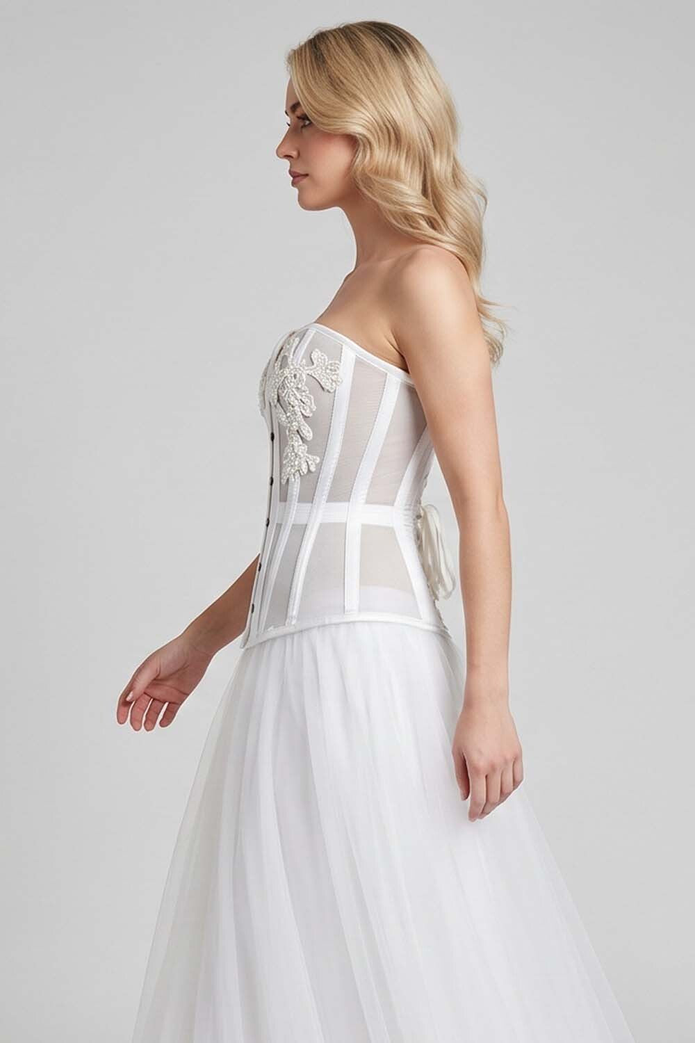 A woman modeling a strapless LUXE NOIR CORSET white wedding dress with structured sheer panels, floral applique on the bodice, and a full tulle skirt against a light gray studio background.