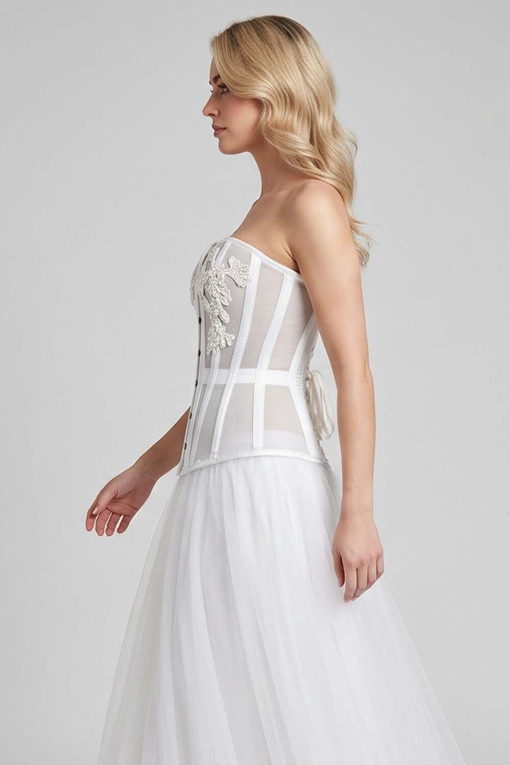 A woman modeling a strapless LUXE NOIR CORSET white wedding dress with structured sheer panels, floral applique on the bodice, and a full tulle skirt against a light gray studio background.